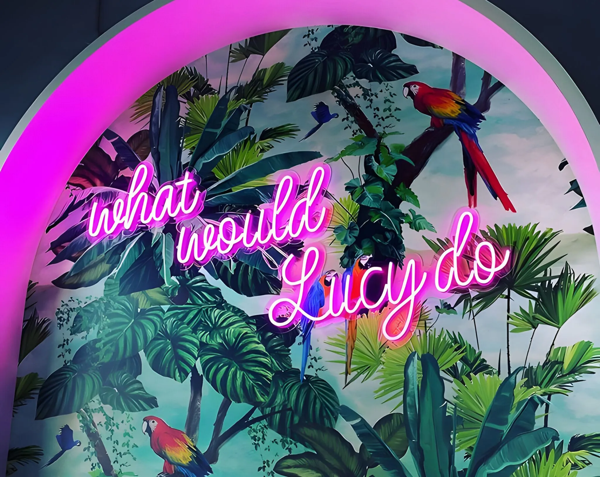 A vibrant mural with lush green tropical foliage and brightly colored parrots, framed by a glowing pink neon light arch featuring the neon-lit phrase "what would Lucy do".