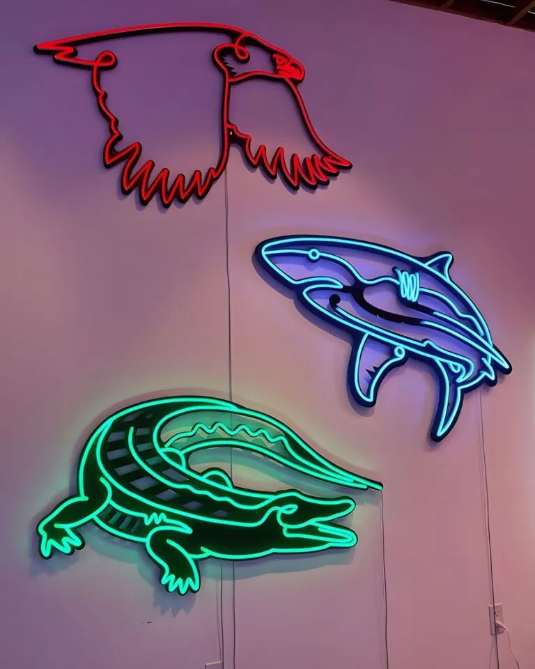 Three neon light sculptures on a wall depicting a red eagle in flight, a blue shark, and a green alligator.