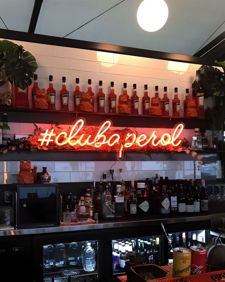 A bar shelf lined with Aperol bottles under a neon sign reading "#clubaperol," with various bottles of alcohol and bar equipment in the foreground.