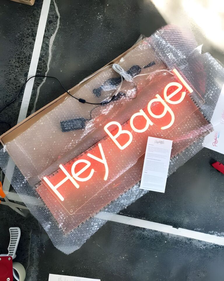A neon sign reading "Hey Bagel" partially wrapped in bubble wrap, with a power cord attached, lying on a marbled floor beside an open cardboard box and scattered papers.