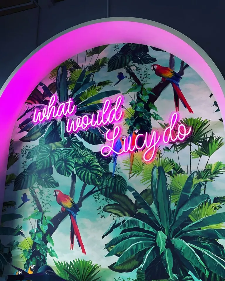 A vibrant neon sign reading "what would Lucy do" against a tropical rainforest mural with parrots and lush foliage, illuminated by a pink glowing arch.