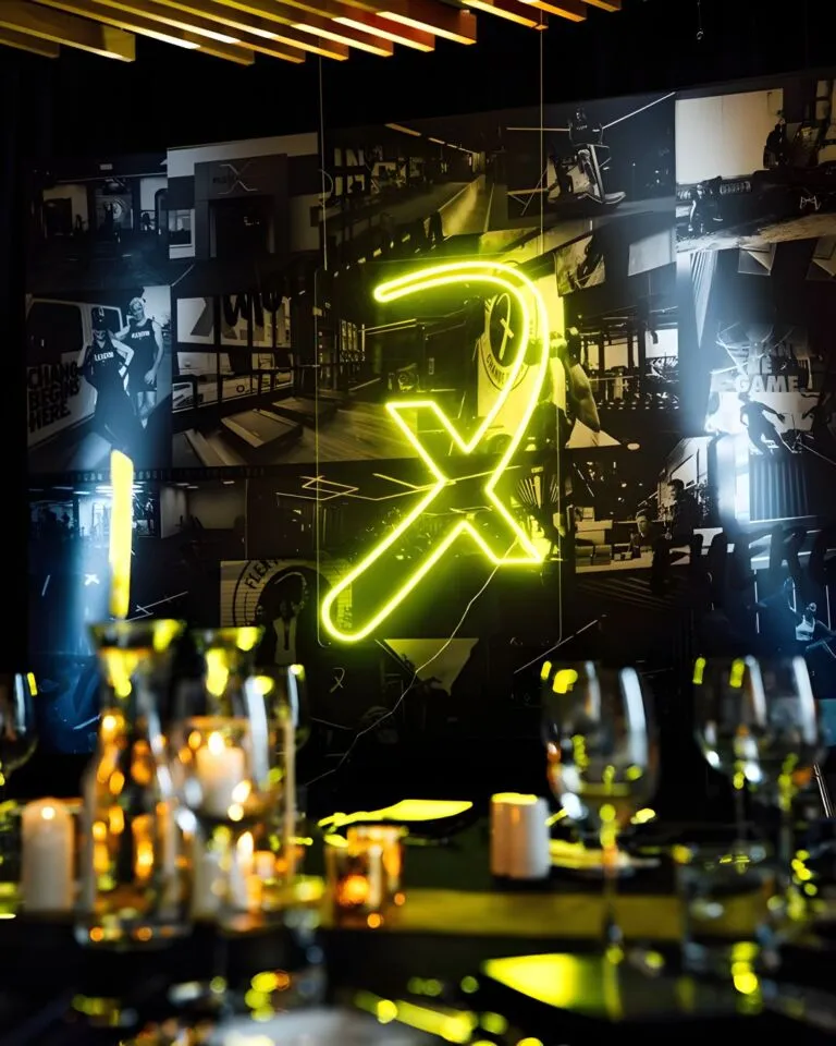 A neon green "X" sign illuminates a modern restaurant interior with empty glasses and candles on tables, casting reflections, with a montage of black and white photos in the background.