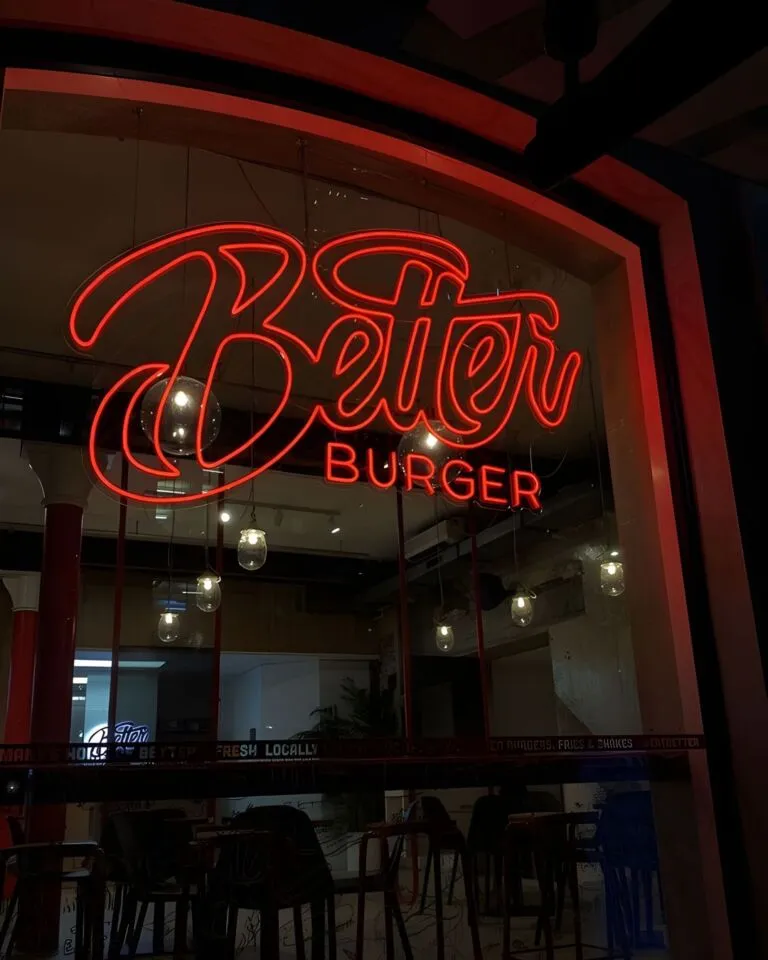 Neon sign of "Better Burger" on a restaurant's window with a dark interior and pendant lights visible, viewed from outside at night.