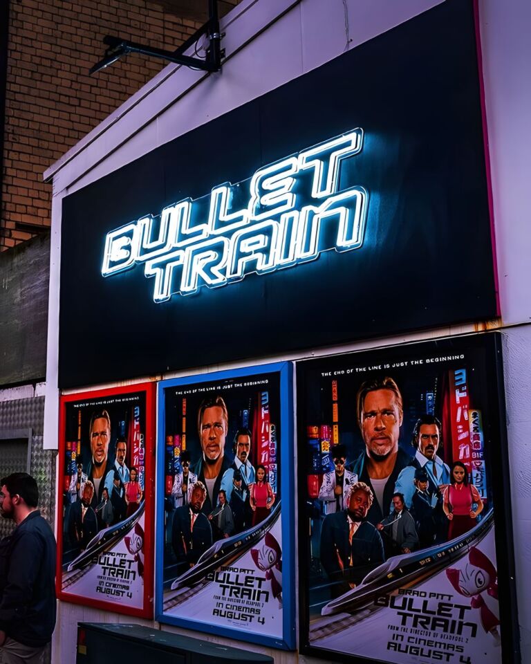 Neon sign reading "BULLET TRAIN" above movie posters with the title "BULLET TRAIN," featuring multiple characters and the release date "IN CINEMAS AUGUST 4." A person is standing to the left, looking at the posters.