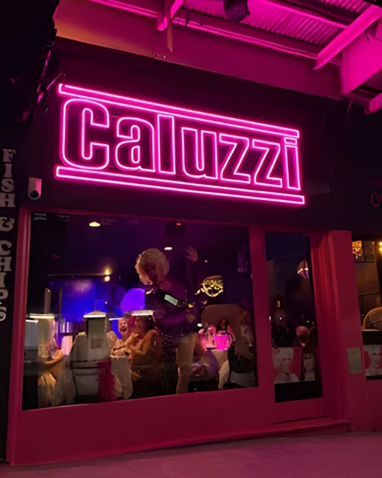 A neon sign that reads "Caluzzi" above a window where people can be seen dining inside the illuminated pink interior of a restaurant.