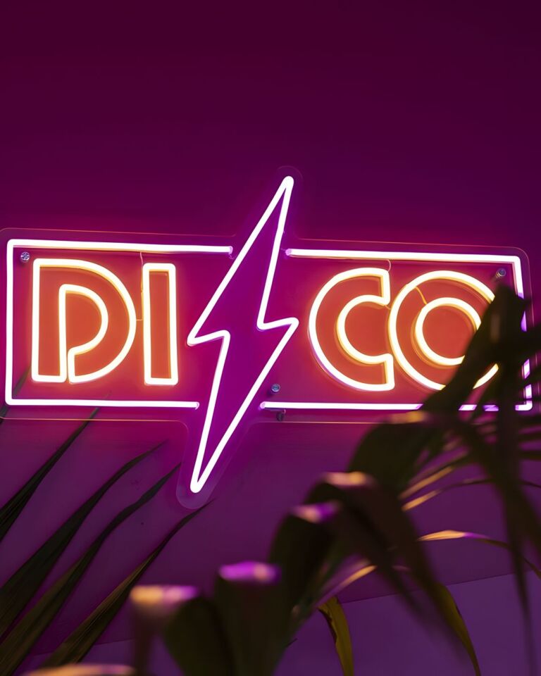Neon sign with the word "DISCO" in capital letters and a lightning bolt symbol, mounted on a purple wall, partially obscured by plant leaves in the foreground.