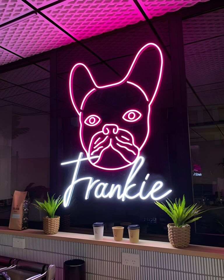Neon sign of a French Bulldog with the name "Frankie" below it, mounted on a window with a pink ceiling light reflection, two potted plants, and three takeaway coffee cups on a counter in the foreground.