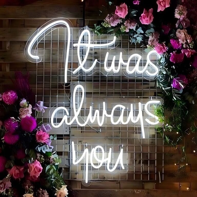 A neon sign on a wooden backdrop with the cursive inscription "It was always you" surrounded by an arrangement of pink and purple flowers.