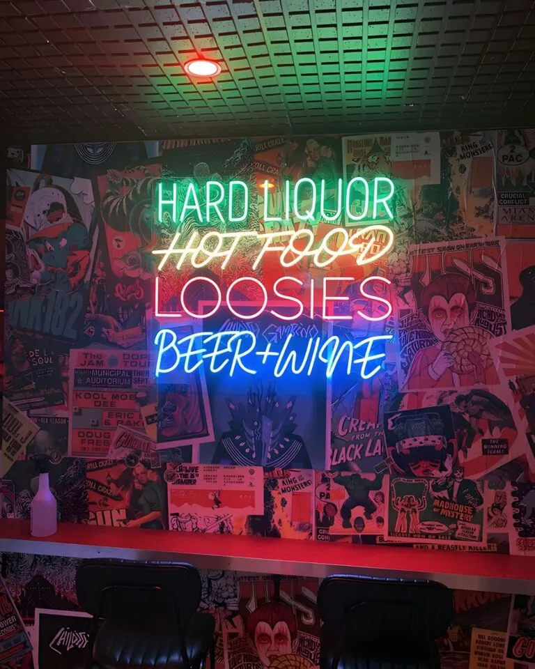 A neon sign with the words "HARD LIQUOR HOT FOOD LOOSIES BEER+WINE" illuminated in bright colors inside a dimly lit bar decorated with a variety of colorful posters and artwork on the walls. Counter seating with black chairs is visible in the foreground.