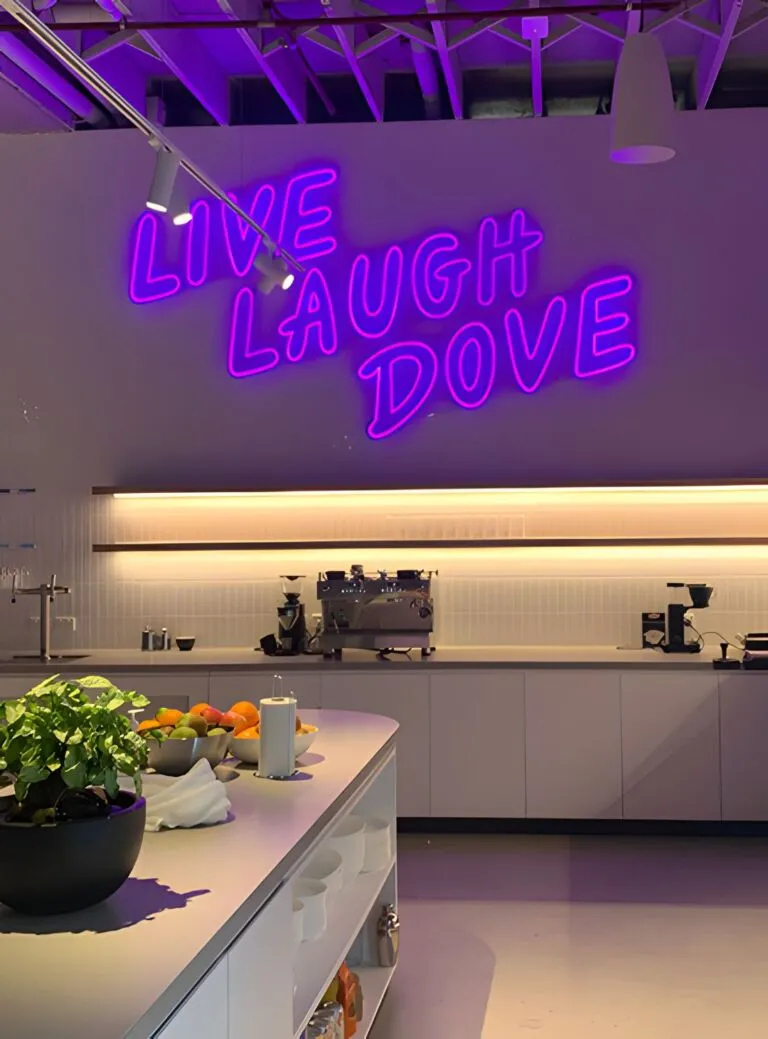A modern kitchen with a neon sign that reads "LIVE LAUGH DOVE" on a mauve wall, illuminated by purple lighting. The counter is adorned with a bowl of citrus fruits, potted plants, and a bottle of milk. A coffee-making station is set up in the background.