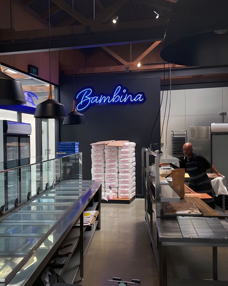A contemporary pizza kitchen with a neon sign reading "Bambina", large pendant lights, a glass display, stacked pizza dough boxes, and a person working in the background.