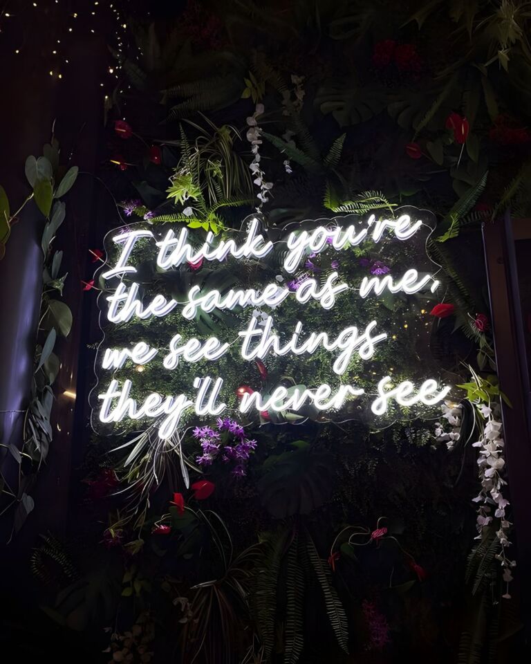 Neon sign with the text "I think you're the same as me, we see things they'll never see" surrounded by a variety of artificial plants and flowers in a dark setting.