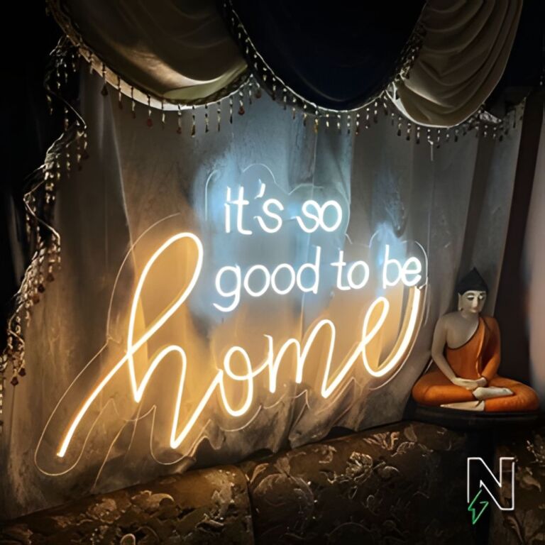 Neon sign reading "It's so good to be home" mounted on a draped fabric background with a statue of a seated Buddha figure to the right, and a dimly lit cozy ambiance.