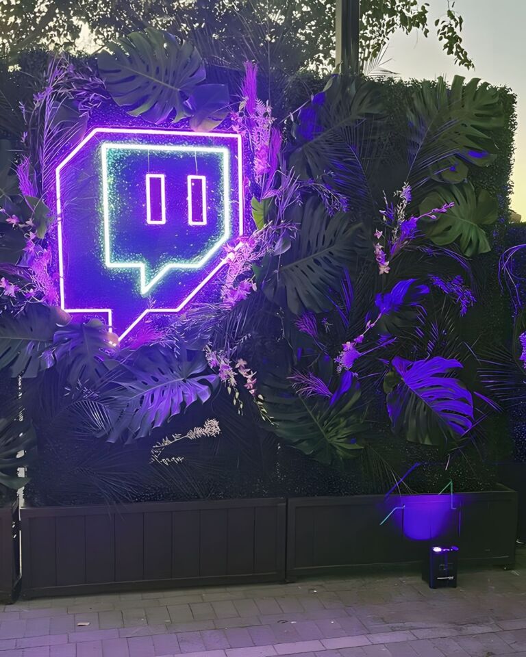 A neon purple Twitch logo illuminated on a feature wall covered with dark green tropical foliage, with a small podium in front of it that is also emitting purple light.